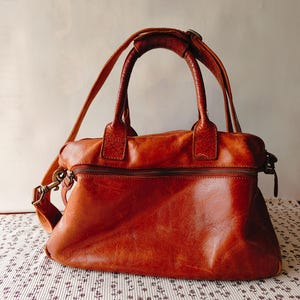 May include: A brown leather handbag with a top handle and a detachable shoulder strap. The bag has a zipper closure and a textured surface. The hardware is a brass color.