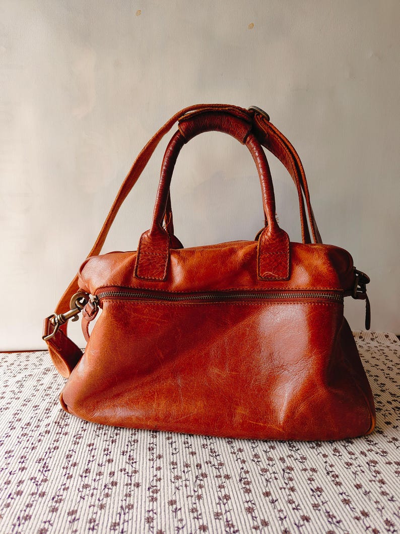 May include: A brown leather handbag with a top handle and a detachable shoulder strap. The bag has a zipper closure and a textured surface. The hardware is a brass color.