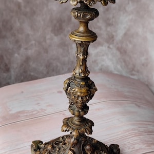 Puede incluir: Candelabro de bronce ornamentado con intrincadas tallas florales y de rostros. El candelabro tiene una base decorativa, una columna central con un rostro esculpido y una copa superior para una vela. Diseño antiguo.