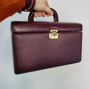 May include: A burgundy vanity case made of leatherette, featuring a gold-coloured clasp and handle fittings. The rectangular case has a textured finish and a curved handle. The case is being held.