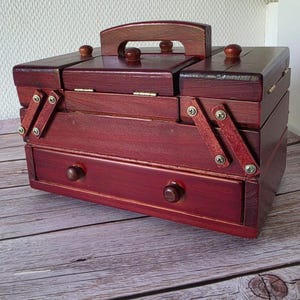 Könnte beinhalten: Ein dunkelrotbrauner Nähkasten aus Holz mit einem mehrstufigen Ausziehmechanismus, einem Tragegriff, aufklappbaren Deckeln und einer unteren Schublade, die mit zwei kleinen, runden Holzknöpfen zum Öffnen versehen ist.