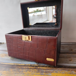 Op de afbeelding: Een vintage, open bruinleren sieradendoosje met een krokodillenleerpatroon. De binnenkant is gevoerd met zwarte stof en heeft een kleine spiegel. De doos heeft een goudkleurige sluiting en scharnieren.