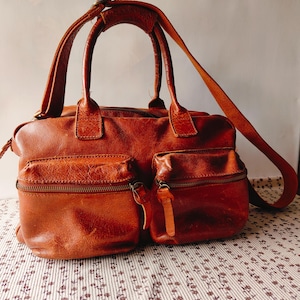 May include: A brown leather satchel bag with two front zippered pockets and a long adjustable strap. The bag has two top handles and a zipper closure. The leather has a distressed appearance.