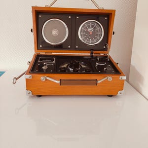 May include: A retro wooden case featuring a speaker, clock, and control panel. The case has a brown handle and silver metal hardware. The clock has a black face with white numerals and hands. The speaker has a silver mesh cover. The case is open, displaying the internal mechanisms.