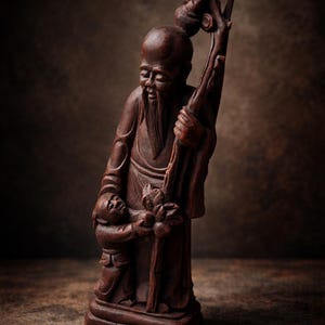 May include: A detailed wooden sculpture of an elderly man with a long beard, holding a staff with a small figure at his feet. The sculpture is a rich brown colour and stands on a rectangular base. The background is a dark, textured surface.