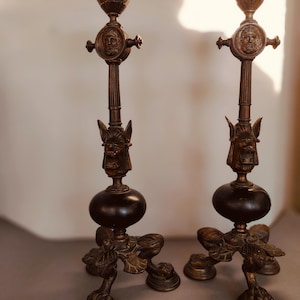 Puede incluir: Dos candelabros antiguos de bronce. Cada uno presenta un diseño detallado con una base redonda, una esfera central y una copa superior. Los candelabros están adornados con caras esculpidas, elementos decorativos y un diseño de pie en forma de garra.
