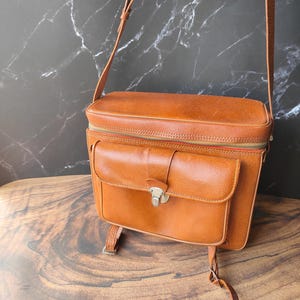 Può includere: Borsa per fotocamera vintage in pelle marrone con tracolla. La borsa presenta una tasca frontale con chiusura in metallo e una chiusura superiore con cerniera. La pelle ha un tono ricco e caldo. La borsa è larga circa 25 cm.