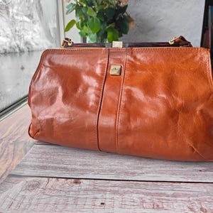 May include: A vintage, rectangular, brown leather handbag with a gold-colored clasp. The bag has a structured design with a central vertical seam and a small gold square emblem. The handle is a dark brown color.