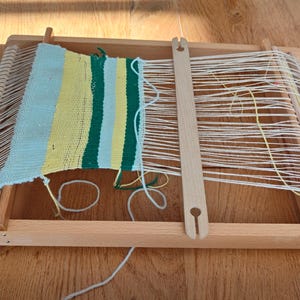Può includere: Un telaio in legno con un pezzo tessuto parzialmente completato. La sezione tessuta presenta strisce blu, gialle e verdi. Fili bianchi e gialli sono tesi sul telaio. Il telaio è su una superficie di legno.