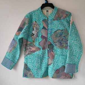 May include: A turquoise quilted jacket with a white and turquoise patterned design. The jacket has a button closure and a mandarin collar. The jacket is decorated with colourful embroidered floral designs.