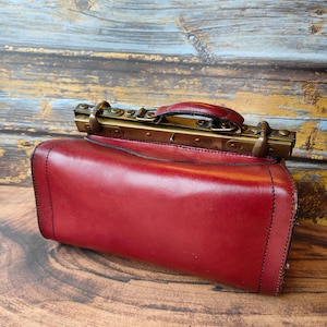 May include: A burgundy leather satchel with a brass handle and clasp. The bag has a rectangular shape with rounded edges and black stitching. The satchel is set against a wooden background.