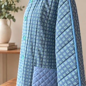 Op de afbeelding: Een doorgestikt jack in blauw- en lichtgroene tinten met een herhalend bloemmotief. Het kledingstuk heeft een hoge kraag, lange mouwen, een knoopsluiting aan de voorzijde met bijpassende knopen en een enkele effen blauwe doorgestikte zak.