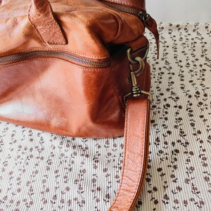 May include: A brown leather handbag with a top handle and a detachable shoulder strap. The bag has a zipper closure and metal hardware. The leather has a distressed look. The bag is sitting on a floral patterned fabric.
