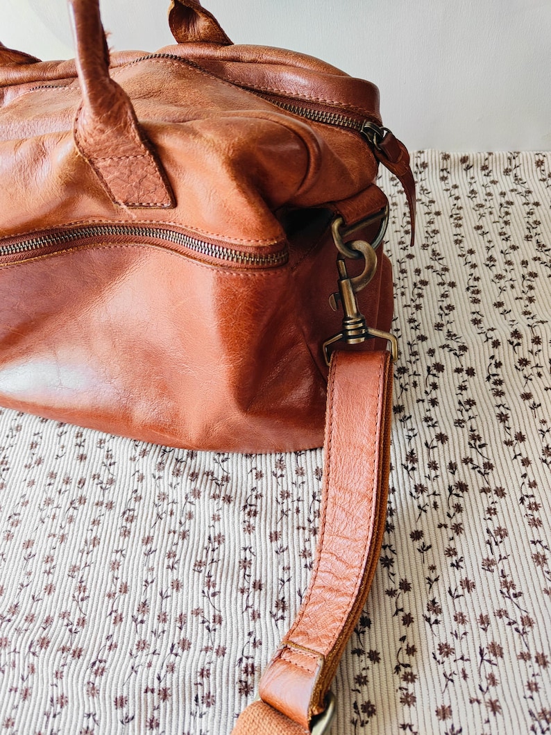 May include: A brown leather handbag with a top handle and a detachable shoulder strap. The bag has a zipper closure and metal hardware. The leather has a distressed look. The bag is sitting on a floral patterned fabric.