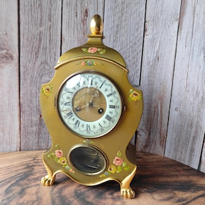 May include: A gold-colored antique clock with a round face featuring Roman numerals and ornate hands. The clock has floral accents and rests on decorative feet. The clock's pendulum is visible through a small, oval window.