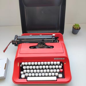 Può includere: Una macchina da scrivere Underwood 319 rossa con tasti bianchi e nastro inchiostro nero. La macchina da scrivere è aperta, rivelando il meccanismo interno. La custodia nera è aperta dietro la macchina. Si vede un piccolo cactus in vaso.