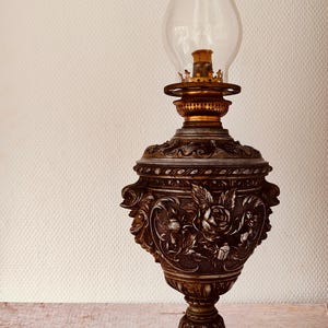 Könnte beinhalten: Eine antike Öllampe mit einem transparenten Glasschornstein und einem verzierten, bronzefarbenen Sockel. Der Sockel weist detaillierte Blumen- und Cherubim-Designs auf. Die Lampe ist ein dekoratives Stück, wahrscheinlich für die Inneneinrichtung.