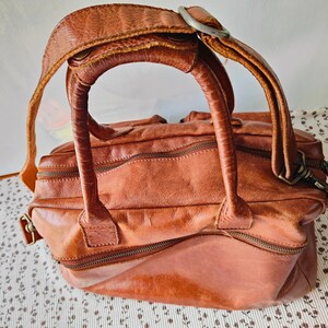 May include: A distressed brown leather handbag with a top handle and a shoulder strap. The bag features multiple zippered compartments and metal hardware. The leather has a worn, textured appearance, suggesting a vintage or rustic style.