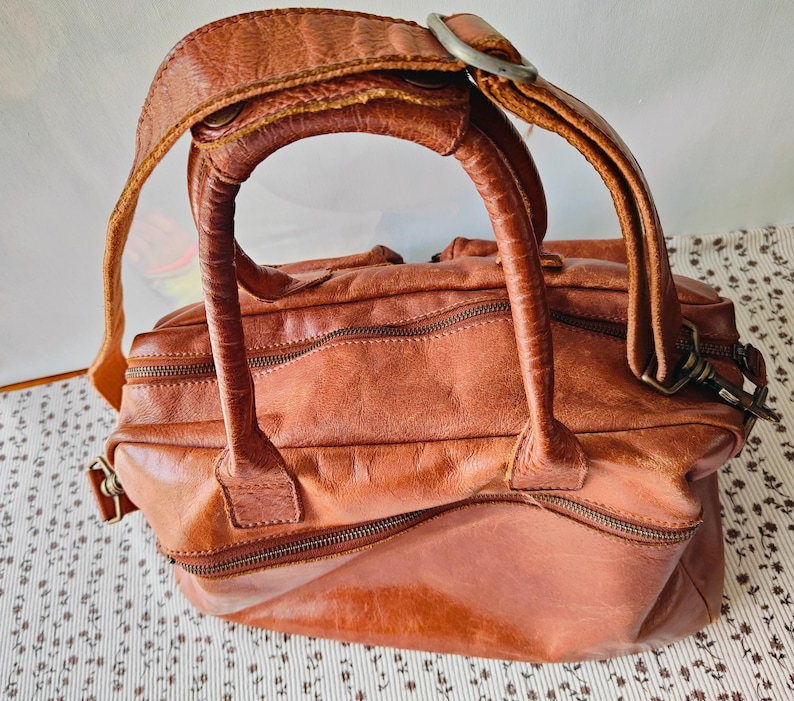 May include: A distressed brown leather handbag with a top handle and a shoulder strap. The bag features multiple zippered compartments and metal hardware. The leather has a worn, textured appearance, suggesting a vintage or rustic style.