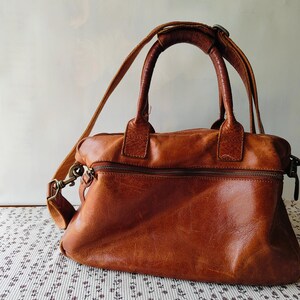 May include: A brown leather handbag with a top handle and a detachable shoulder strap. The bag has a zippered top closure and a front zippered pocket. The leather has a distressed look. The bag is sitting on a floral patterned surface.
