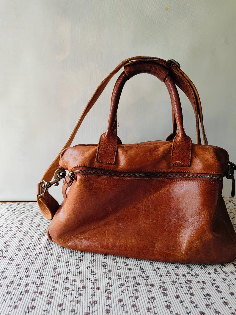 May include: A brown leather handbag with a top handle and a detachable shoulder strap. The bag has a zippered top closure and a front zippered pocket. The leather has a distressed look. The bag is sitting on a floral patterned surface.