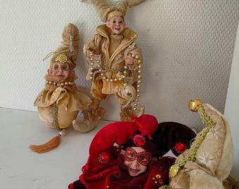 May include: Several decorative jester figurines in various poses. The jesters wear elaborate costumes in gold, red, and brown, with beaded accents and pointed hats. One jester is standing, while others are seated or lying down.