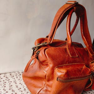 May include: A distressed, brown leather satchel bag with a top handle and a shoulder strap. The bag features a front pocket with a zipper and a brass-colored clasp. The leather has a worn, vintage appearance.