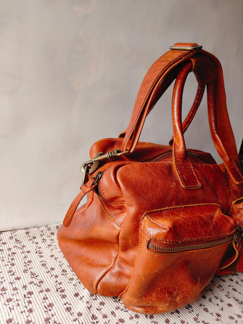 May include: A distressed, brown leather satchel bag with a top handle and a shoulder strap. The bag features a front pocket with a zipper and a brass-colored clasp. The leather has a worn, vintage appearance.