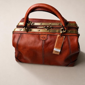 May include: A cognac-colored leather doctor-style bag featuring a structured top handle, brass-toned metal hardware accents along the top opening, a zippered closure, and a small leather tag with the text 1150-03 and 133 printed on the front.