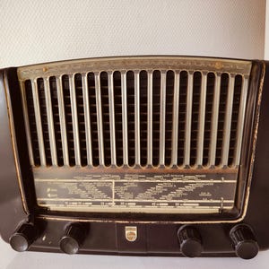 Puede incluir: Radio negra vintage con rejilla plateada y cuatro perillas negras. La radio tiene un dial con marcas de estaciones y un logotipo de Philips. El diseño de la radio es de una época pasada.