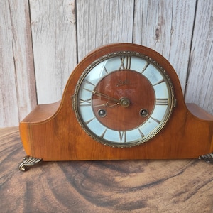 Op de afbeelding: Een vintage houten mantelklok met een lichtbruine afwerking. De klok heeft Romeinse cijfers en een messingkleurige rand. De klok heeft decoratieve voetjes en een gebogen bovenkant.