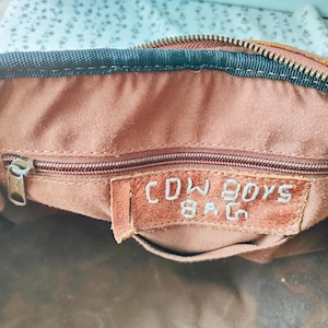 May include: Close-up of the interior of a brown leather bag. The bag has a zipper closure and a small leather tag that reads "COWBOYS BAG" in embossed lettering. The interior fabric is a lighter brown color.