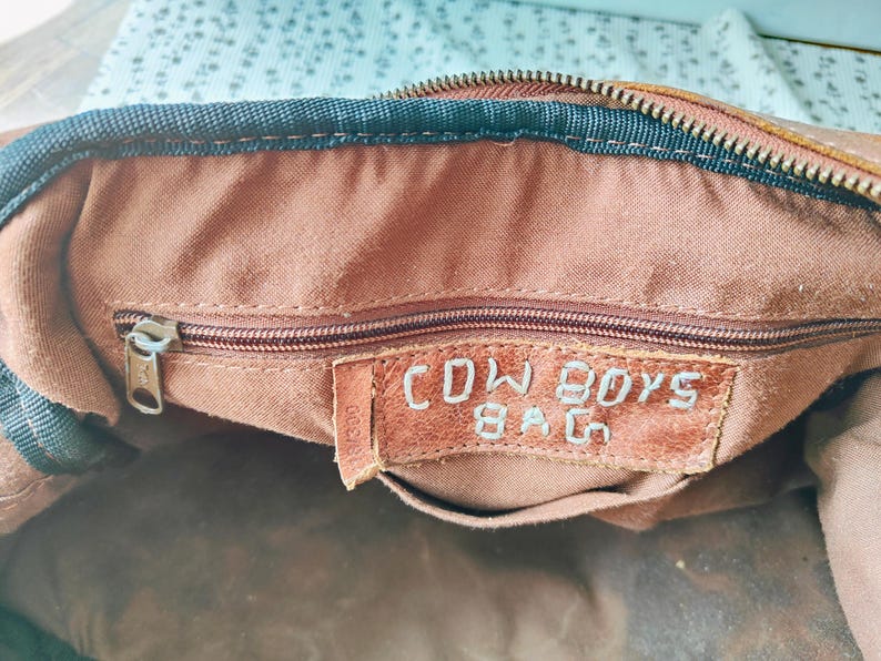 May include: Close-up of the interior of a brown leather bag. The bag has a zipper closure and a small leather tag that reads "COWBOYS BAG" in embossed lettering. The interior fabric is a lighter brown color.