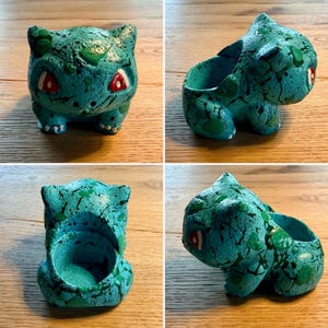 Könnte beinhalten: Ein tealfarbener Bulbasaur-Pflanztopf mit roten Augen und grünen Akzenten. Der Keramik-Pflanztopf hat eine strukturierte Oberfläche und ist so gestaltet, dass er der beliebten Pokémon-Figur ähnelt. Der Pflanztopf eignet sich für kleine Pflanzen oder Sukkulenten.