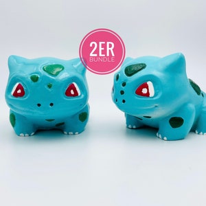 Könnte beinhalten: Zwei handbemalte Keramikfiguren eines Pokemon-Charakters, Bisasam. Die Figuren sind etwa 7,5 cm groß und Teil eines 2ER-Bundles.