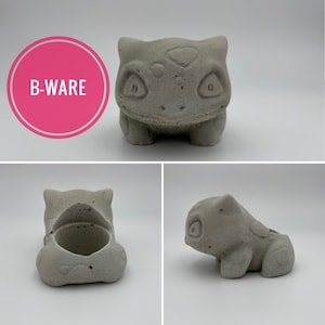 Könnte beinhalten: Ein grauer Beton-Pflanztopf in Form einer Zeichentrickfigur. Der Pflanztopf hat eine runde Öffnung für Pflanzen und ist in Form eines Bulbasaur. Der Text "B-WARE" befindet sich in einem rosa Kreis.