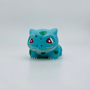 Könnte beinhalten: Hellblaue Bulbasaur-Figur mit grünen Flecken und Akzenten. Die Figur hat rote Augen und einen lächelnden Ausdruck. Die Figur ist ein beliebter Charakter aus dem Pokémon-Franchise.