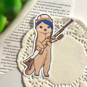 Peut inclure: Un autocollant représentant une belette de dessin animé portant un chapeau de marin bleu et blanc et tenant un fusil. La belette est brune avec un museau brun clair et porte un nœud bleu sur la tête.