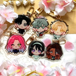 Op de afbeelding: Collectie van zes sleutelhangers met anime-stijl karakters met goudkleurige sluitingen. Elke sleutelhanger toont een ander karakter met unieke haarkleur en outfits. De sleutelhangers zijn omringd door roze en witte bloemen.