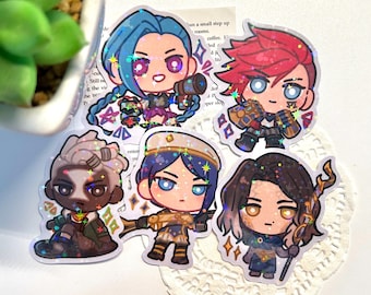 May include: Five holographic stickers featuring cartoon characters from the video game League of Legends. The characters are Jinx, Vi, Jayce, Caitlyn, and Ekko. The stickers are on a white background.