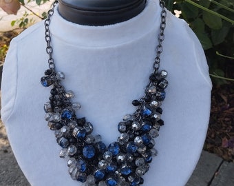 May include: A silver chain necklace with a cascading cluster of blue and clear glass beads. The necklace is displayed on a white mannequin.