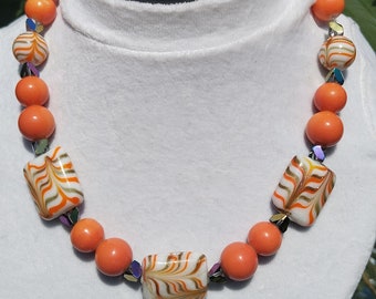 May include: A necklace made of orange and white beads. The beads are round and have a swirl pattern. The necklace is on a white mannequin.