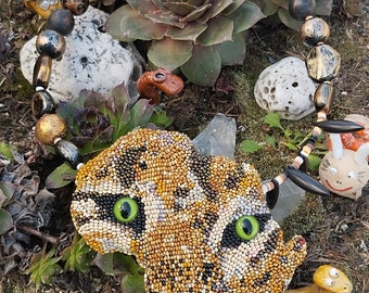 May include: A beaded tiger face in the shape of Africa, with green eyes, surrounded by a black and gold beaded necklace. The necklace is on a bed of moss and rocks.