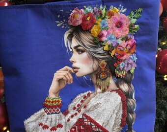 May include: A blue tote bag featuring a colourful floral design and a woman's face. The woman is wearing a white embroidered shirt with red accents and a beaded flower crown.