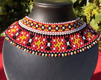 May include: A red, white, black and yellow beaded choker necklace with a geometric pattern. The necklace is displayed on a black velvet stand.