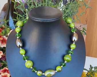 May include: A green beaded necklace on a black display bust. The necklace has large, oval, and round green beads, with smaller green and silver beads. The background includes flowers and a wine bottle.