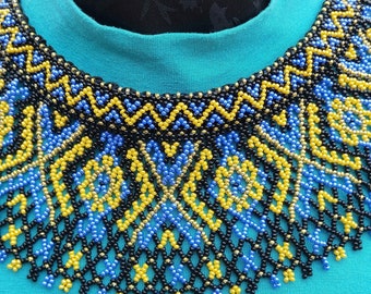 May include: A beaded necklace with a geometric pattern in black, blue and yellow. The necklace is on a turquoise shirt.