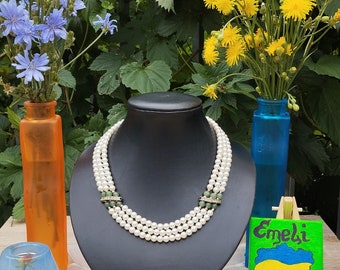 May include: A three-strand pearl necklace with green gemstone accents on a black display stand. The necklace is surrounded by blue and yellow flowers in vases.