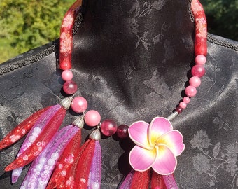 May include: A pink and red beaded necklace with a pink flower charm and red and purple feather-like tassels. The necklace is on a black mannequin.