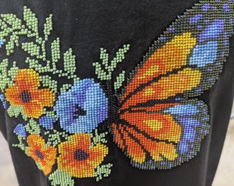 May include: A black t-shirt featuring a beaded butterfly and floral design. The butterfly is orange, yellow, and blue, and the flowers are orange, yellow, and blue.